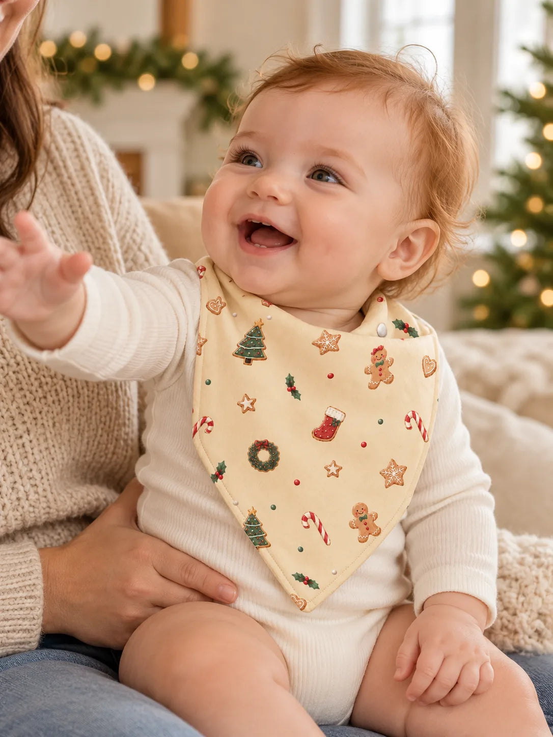 Baby on a parent’s lap wearing the MossiBaby Christmas Joy bandana bib and reaching upward beside a Christmas tree.