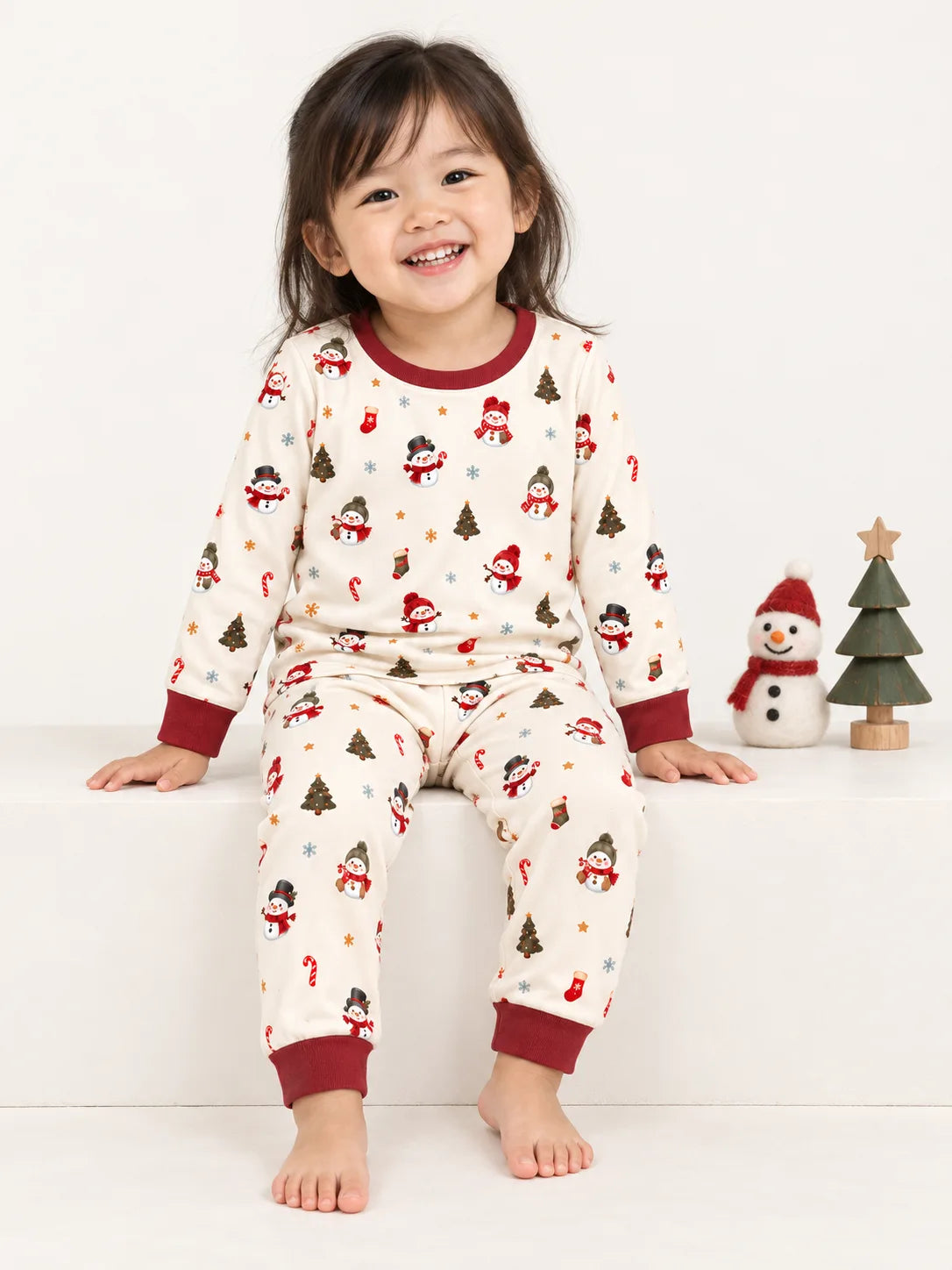 Toddler sitting in a studio wearing Mossibaby Christmas Snowman ivory cream bamboo long sleeve pajama set with cranberry red trim