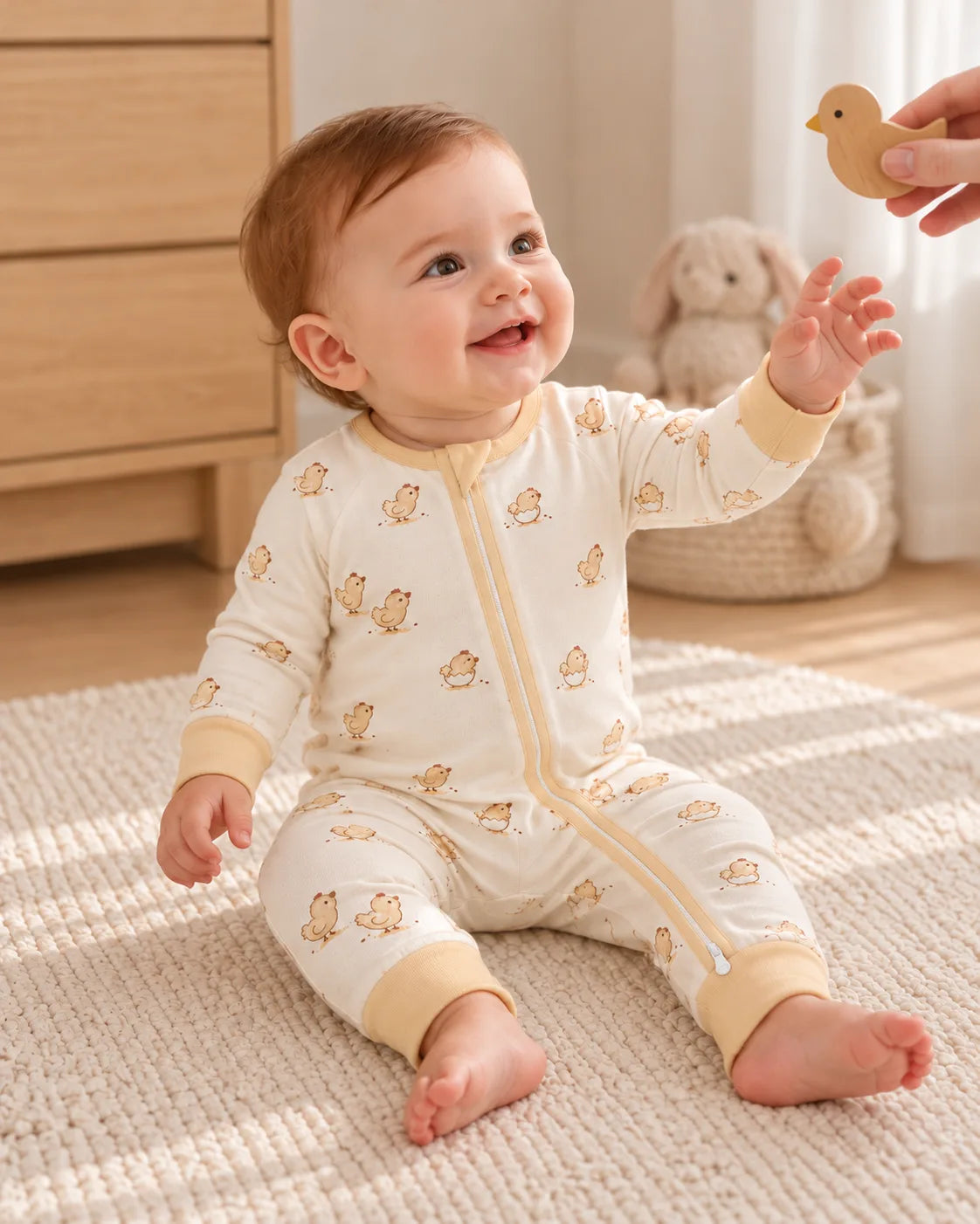 Little Hatchlings Long Zipper Romper. Baby seated beside a nursery dresser with a chick toy held by an adult.