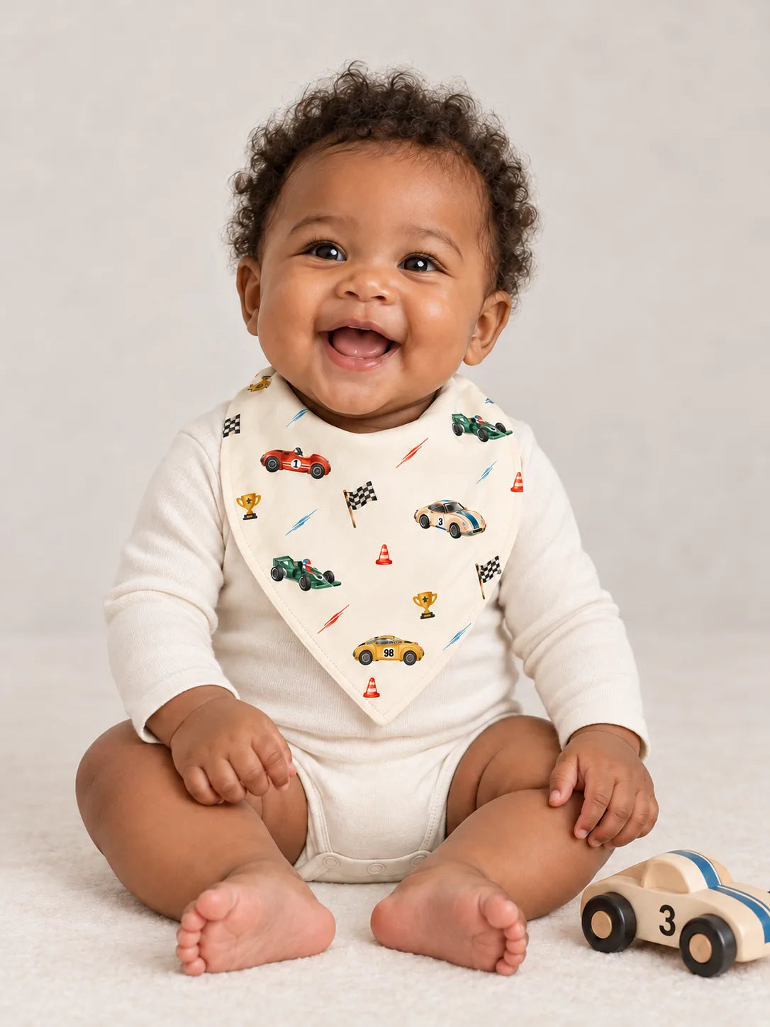 Smiling baby wearing the Vintage Racers bamboo bandana bib over an ivory bodysuit in a studio.
