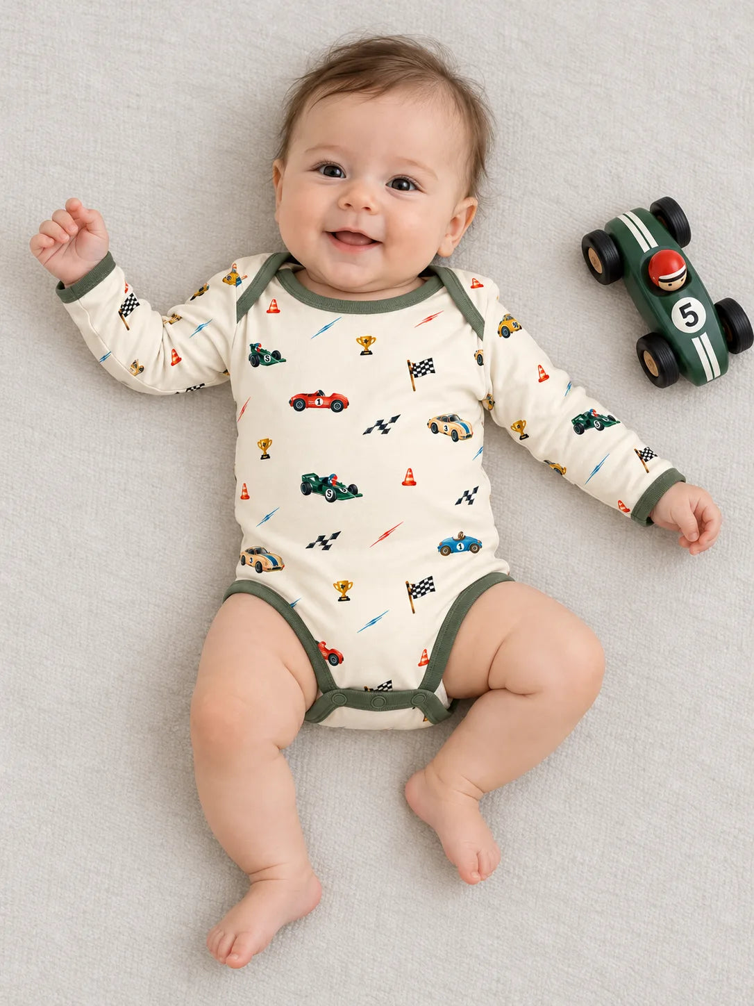 Smiling baby lying in the Vintage Racers bamboo long sleeve bodysuit beside a toy racing car in a studio.