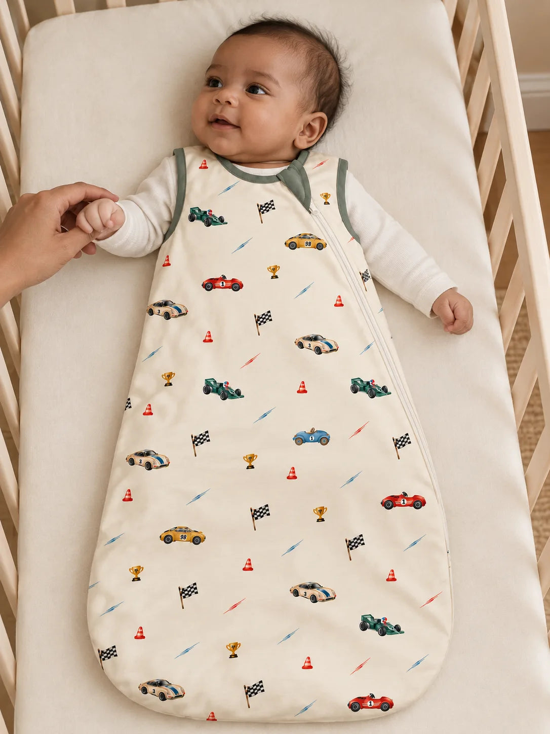 Baby lying in the Vintage Racers bamboo sleep bag on a cot mattress while holding a parent's hand.