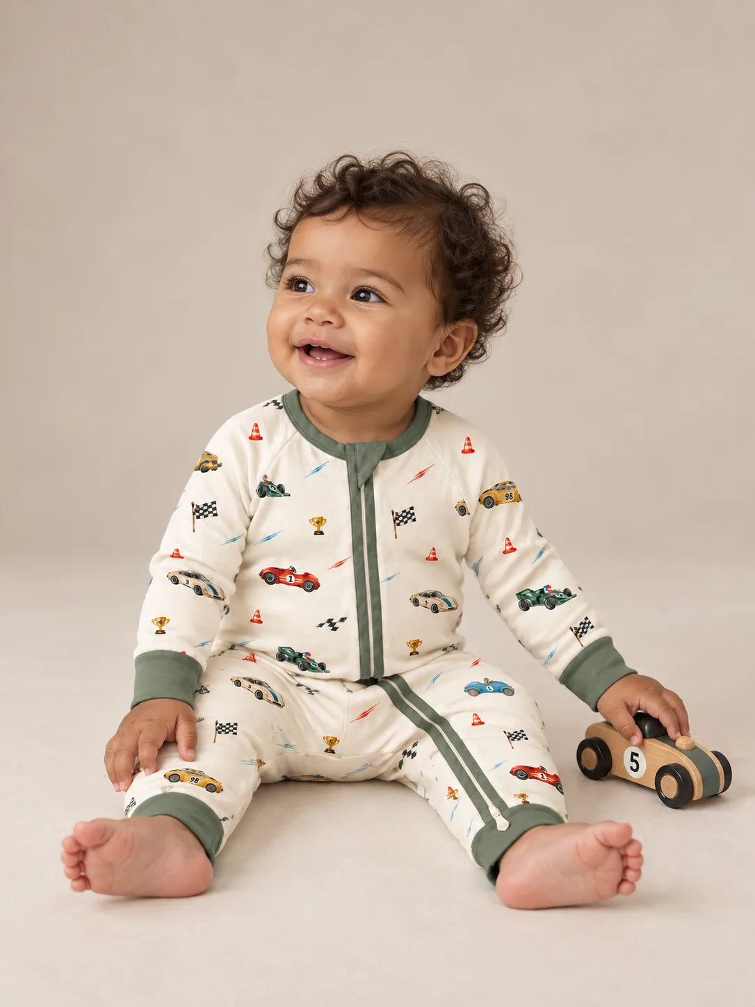 Baby sitting in the Vintage Racers bamboo zipper romper beside a wooden racing car in a studio.