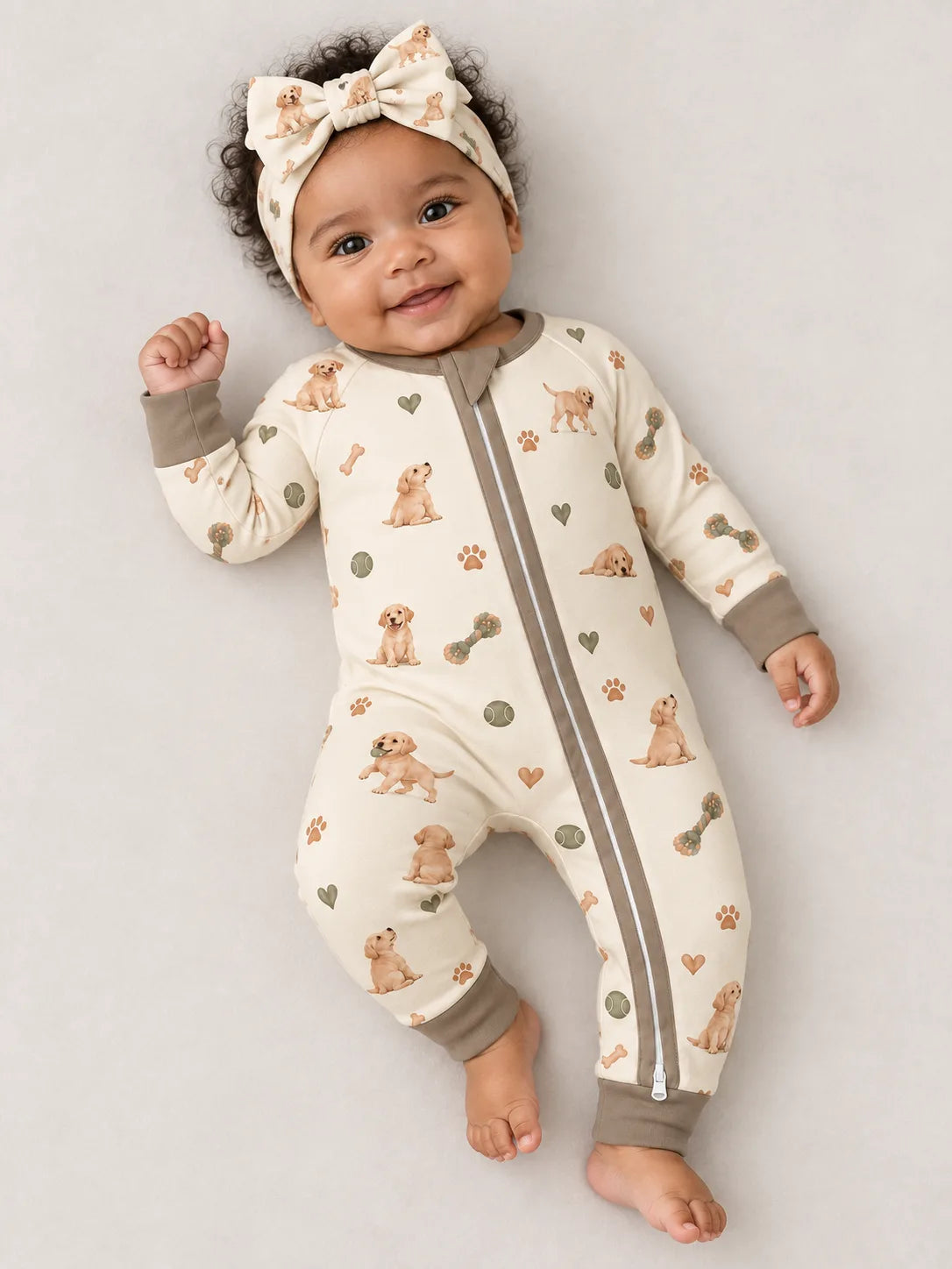 Baby wearing the MossiBaby Little Labradors bow headband with a matching zipper romper against a pale studio backdrop